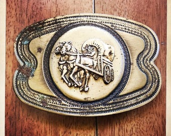 Könnte beinhalten: Eine vintage Gürtelschnalle aus Messing mit einem verzierten Design. Die Schnalle zeigt ein Reliefbild von zwei Pferden, die einen Wagen ziehen.
