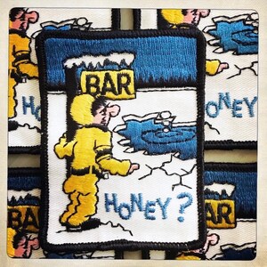 May include: Embroidered patch featuring a cartoon character in a yellow snowsuit standing in a snowy landscape with a sign that reads "BAR" and the text "Honey?"