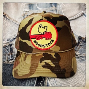 Puede incluir: Una gorra de camionero de camuflaje con un parche bordado amarillo y rojo que presenta un signo de paz y la palabra "Woodstock".