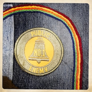 May include: A close-up of a Public Enemy No. 1 patch on a denim jacket. The patch is a yellow circle with a bell inside and the words "Public Enemy No. 1" around the outside. The jacket has a rainbow-colored stripe sewn on the side.