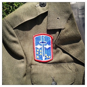Könnte beinhalten: Eine grüne Militärjacke mit einem roten, weißen und blauen Aufnäher mit einem Schwert und Sternen. Der Aufnäher ist auf der linken Seite der Jacke angenäht.