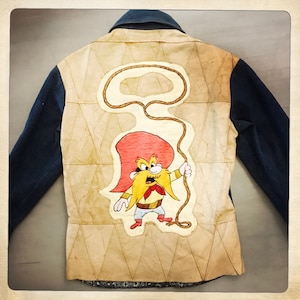 May include: A denim jacket with a brown suede back panel featuring a cartoon character with a red hat, yellow beard, and a lasso. The character is wearing a red bandana and brown pants.