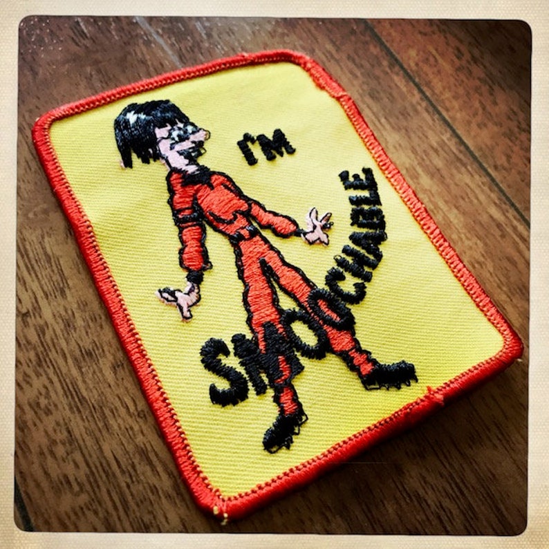 May include: A yellow and red embroidered patch with a cartoon figure in a red jumpsuit and the text "I'm Snackable".