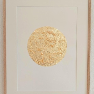 Könnte beinhalten: Ein gerahmtes Kunstwerk mit einem zentralen Kreis aus Blattgold, der eine texturierte, zerknitterte Oberfläche aufweist. Das Kunstwerk ist vor einem schlichten weißen Hintergrund in einem hellen Holzrahmen platziert und wirkt klar.