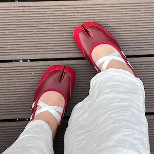 May include: A pair of deep red split-toe shoes featuring a smooth leather-like vamp and suede-textured heel and toe caps. The shoes have a low-cut silhouette with white crisscross elastic straps across the instep, worn with white trousers.