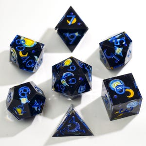 Pode incluir: Um conjunto de sete dados poliédricos com uma base de resina azul-escura e preta, com motivos incrustados de luas crescentes e estrelas amarelas. Cada dado tem gravações numéricas em azul brilhante, incluindo um d20, d12, d10, d8, d6, d4 e um dado de percentagem.