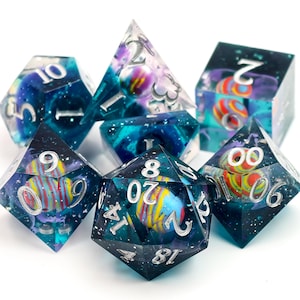 Puede incluir: Un juego de siete dados poliédricos hechos de resina translúcida azul oscuro y morada con purpurina incrustada y esferas internas de rayas multicolores. Cada dado presenta números grabados de color plateado, incluido un dado de veinte caras.