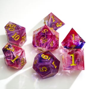 Pode incluir: Um conjunto de sete dados poliédricos feitos de resina translúcida com uma mistura rodopiante de cores rosa, roxa e branca. Cada dado apresenta números gravados preenchidos com tinta dourada, incluindo um d20, d12, d10, d8, d6, d4 e um dado percentual.