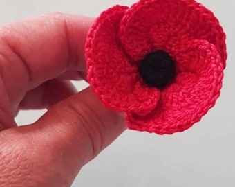 May include: A handmade, crocheted red poppy flower with a black center. The flower is in full bloom, showcasing detailed stitching and a textured appearance. The poppy is a symbol of remembrance.