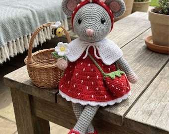 May include: A handmade crochet mouse doll wearing a red strawberry dress, matching shoes, and a red polka dot headband. The doll has a white collar, a green strawberry purse, and is holding flowers. A small basket with a bee is next to the doll.