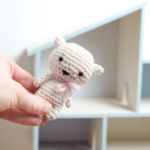 May include: A small, crocheted off-white bear with black eyes and a small black stitched mouth. The bear has small ears and a pink bow around its neck. The bear is held in a hand, with a dollhouse in the background.