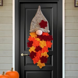 Könnte beinhalten: Handgemachte Herbst-Wichtel-Türdekoration. Der Wichtel hat eine gehäkelte graue Mütze mit rotem Blätterakzent, eine rosa Nase und einen Bart aus bunten gehäkelten Blättern in Orange-, Gelb- und Rottönen. Der Wichtel hängt an einer schwarzen Tür.
