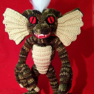 Puede incluir: Criatura de ganchillo con cuerpo de camuflaje, alas color canela y ojos rojos. El peluche hecho a mano tiene un pecho blanco y un mechón de pelo blanco. La criatura tiene una amplia sonrisa con labios rojos y dientes blancos.