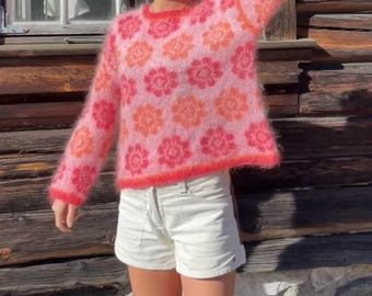May include: A pink, fuzzy sweater with a floral pattern in shades of pink and orange. The sweater has a crew neck and long sleeves. The person is wearing white shorts. The background is a wooden wall.