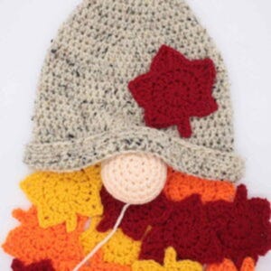 May include: A handmade gnome with a speckled gray hat, a red leaf accent, and a peach-colored nose. The gnome's beard is made of crocheted leaves in shades of yellow, orange, and red, perfect for fall decor.