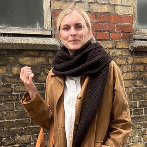 May include: A woman wearing a dark brown knitted scarf, a tan coat, and a white button-down shirt. The background is a brick wall and a window.