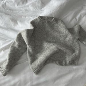 Peut inclure: Un pull en tricot gris clair avec un col rond et des manches longues. Le pull est fait d'un fil texturé et a des poignets et un ourlet côtelés. Le vêtement est posé sur une surface blanche et rayée.