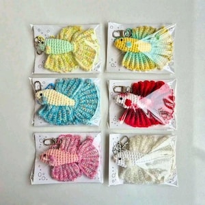 Puede incluir: Seis llaveros de crochet hechos a mano con forma de pez betta, envasados individualmente en plástico transparente. Cada llavero presenta una combinación de colores única, incluyendo amarillo, azul, rojo, rosa y blanco, con aletas y cuerpos detallados. Los llaveros están sujetos a cierres metálicos plateados.