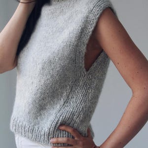 May include: A light gray, sleeveless knit sweater with a high neck. The sweater has a ribbed hem and a textured appearance. The garment is made of a soft, possibly wool, material. The style is a casual, everyday wear.