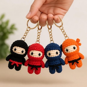 May include: Four crochet ninja keychains featuring small figures in black, red, navy blue, and orange yarn. Each ninja has a beige face, black bead eyes, and a tied belt. The figures are attached to gold-toned metal key rings with short chains.
