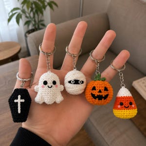 May include: Five crochet keychains featuring Halloween themes: a black coffin with a white cross, a white ghost with pink cheeks, a white mummy, an orange jack-o'-lantern with a green stem, and a yellow, orange, and white candy corn piece.