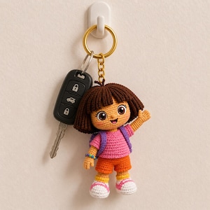 Może przedstawiać: Szydełkowy brelok do kluczy Dora the Explorer ze złotym kółkiem. Lalka ma na sobie różową koszulkę, pomarańczowe szorty i fioletowy plecak. Brelok jest przymocowany do kluczyka do samochodu.