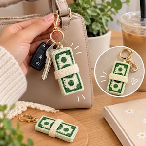 May include: A rectangular crochet keychain designed to resemble a stack of green and cream-colored paper money, secured with a cream-colored band. The keychain features a gold-toned metal lobster clasp and ring, shown attached to keys and a handbag.
