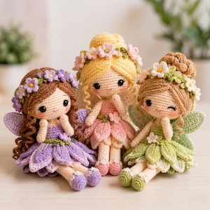 Op de afbeelding: Drie handgemaakte gehaakte feeënpoppen. Elke pop heeft een bloemenkrans, vleugels en een rok in de vorm van een bloemblad. De poppen hebben verschillende haarkleuren: bruin, blond en lichtbruin. De rokken zijn paars, roze en groen. De poppen zijn ongeveer 15 cm hoog.