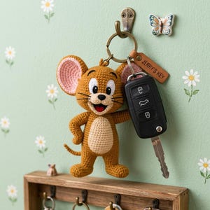 Può includere: Portachiavi di Jerry Mouse all'uncinetto con una chiave dell'auto. Il topo marrone ha orecchie rosa e una pancia beige. Un'etichetta in pelle marrone recita "JERRY'S KEY". Il portachiavi è appeso a una parete con decorazioni di margherite e farfalle.