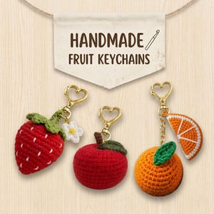 May include: Three handmade crochet fruit keychains with gold heart-shaped clasps. The set includes a red strawberry with a white flower, a red apple with a green leaf, and an orange with a green leaf and a slice of orange attached by a chain.