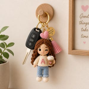 Op de afbeelding: Handgemaakte gehaakte poppen sleutelhanger met bruin haar, een blauwe jeans en een witte trui met de letter "A". De pop houdt een koffiekopje vast en heeft een roze hart en kwast. De sleutelhanger is bevestigd aan een autosleutel.