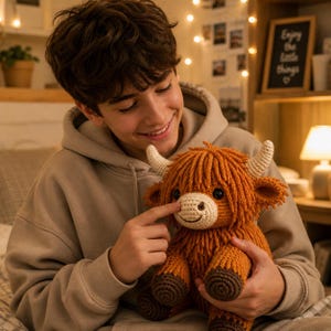 May include: A handmade crochet Highland cow plush toy. The plush features a shaggy orange body, cream-colored horns, and a smiling face. The cow is held by a person wearing a tan hoodie.