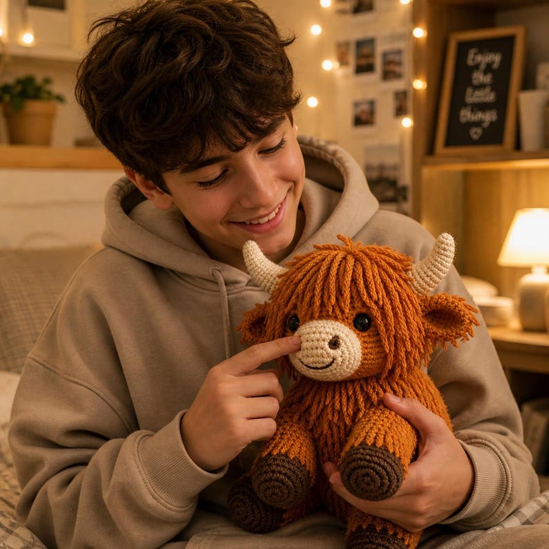 May include: A handmade crochet Highland cow plush toy. The plush features a shaggy orange body, cream-colored horns, and a smiling face. The cow is held by a person wearing a tan hoodie.