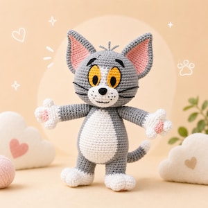 May include: A crocheted Tom cat doll in gray, white, and pink. The cat has large yellow eyes, black whiskers, and a white belly. Its arms are outstretched, and it has a small tail. The doll is handmade and stands on a light-colored surface.
