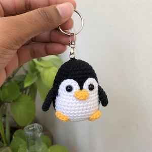 May include: A small, round, handmade crochet penguin keychain featuring black yarn on the head and back, white yarn on the belly and face, two black plastic bead eyes, and a small orange yarn beak and feet, attached to a silver metal keyring.