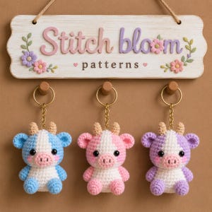 May include: Three crochet cow keychains in blue, pink, and purple, with white faces and accents. They hang below a wooden sign that reads "Stitch bloom patterns" in pastel letters, with floral embellishments.