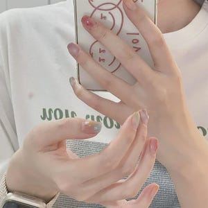 Puede incluir: Una camiseta blanca con texto verde y una funda de smartphone con un diseño de cara sonriente en líneas rojas. Las uñas están pintadas con un esmalte degradado multicolor en tonos azul, amarillo y rosa, creando un efecto suave y difuminado.