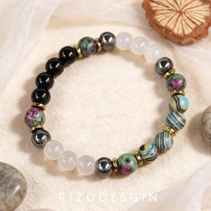May include: A beaded bracelet featuring a mix of round black onyx, white quartz, metallic hematite, green and pink ruby zoisite, and blue and green striped malachite beads, accented with small gold-toned metal spacers throughout the design.