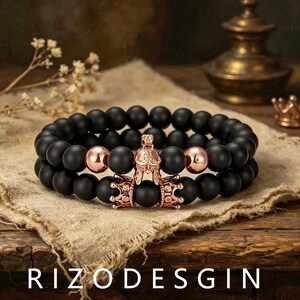 May include: A double-strand bracelet featuring matte black spherical beads, two polished rose gold-tone spherical beads, a rose gold-tone helmet charm, and a rose gold-tone crown charm with small clear stones. The text RIZODESGIN is visible.