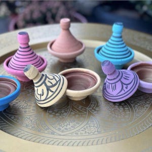Può includere: Una collezione di piccole e colorate pentole tagine in rosa, blu, viola e beige. I tagine hanno coperchi decorativi e sono su un vassoio dorato con un disegno inciso. Sono tradizionali pentole da cucina marocchine.