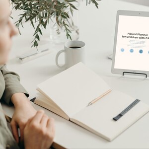 May include: A white tablet displaying "Parent Planner for Children with CAPD" sits on a white desk. An open notebook with a pencil and a black and silver pen holder are in front of the tablet. A white mug and a vase with greenery are also on the desk.