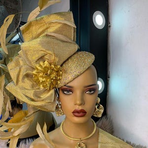 Può includere: Un cappello dorato e ornato con un grande fiocco e decorazioni floreali. Il cappello è abbinato a orecchini e collana dorati. Il manichino ha un trucco occhi scuro e rossetto rosso. Un accessorio di moda di grande effetto.