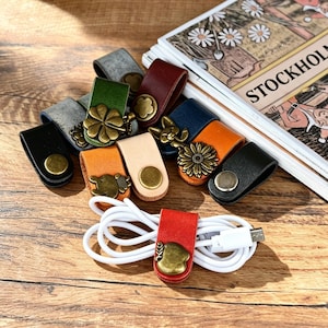 Op de afbeelding: Een verzameling lederen kabelorganizers in diverse kleuren zoals zwart, grijs, groen, beige, blauw, oranje en rood. Elke band is voorzien van een metalen drukknoop met decoratieve vormen zoals bloemen, klavertjes en appels.