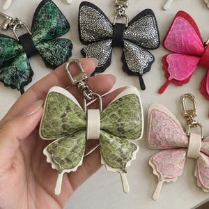May include: A collection of eight butterfly-shaped keychains featuring various textures and colours, including snakeskin patterns in black, white, green, and pink, as well as metallic blue, iridescent, and silver finishes, each with a metal clasp.