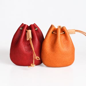 May include: Two small drawstring pouches made of textured leather. One pouch is deep red with a subtle embossed four-leaf clover design, and the other is a bright orange. Both feature light tan leather cords threaded through metal eyelets.