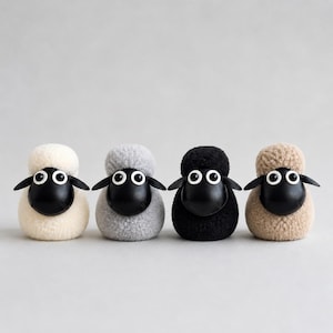 May include: Four small sheep figurines arranged in a row. Each sheep has a textured, wool-like body in cream, light gray, black, or tan, paired with a smooth, matte black head, two black ears, and two large, round white eyes with black pupils.