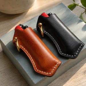 May include: Two leather lighter cases, one in tan and one in black, each featuring white contrast stitching, a brass-toned metal ring attachment, and a contoured shape that holds a red and black lighter securely within the protective sleeve.