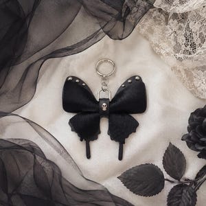 May include: Black leather butterfly keychain with silver rivets and a small skull charm. The keychain has a silver ring and is set against a backdrop of black fabric, lace, and a black rose.