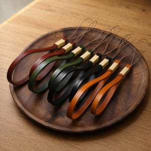 Puede incluir: Una colección de seis correas de muñeca con bucles de cuero de colores en tonos rojo oscuro, verde bosque, negro y tostado. Cada correa está asegurada con un conector rectangular de color claro e incluye un bucle de cordón fino y flexible.