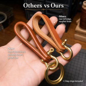 May include: Two brown leather key loops with brass hardware and key rings. The text reads Others vs Ours, Others: raw cut edge, rougher finish, Ours: hand-burnished, smooth sheen, great feel, and + 2 key rings included.